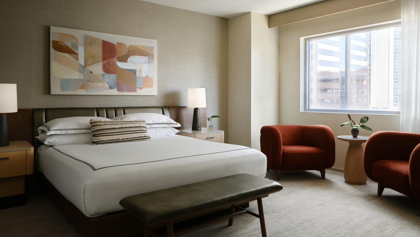 Modern hotel bedroom with a large bed, abstract art, two burnt orange armchairs, and a city view window