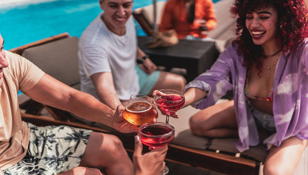 Friends clink their cocktail glasses as they sit by a pool